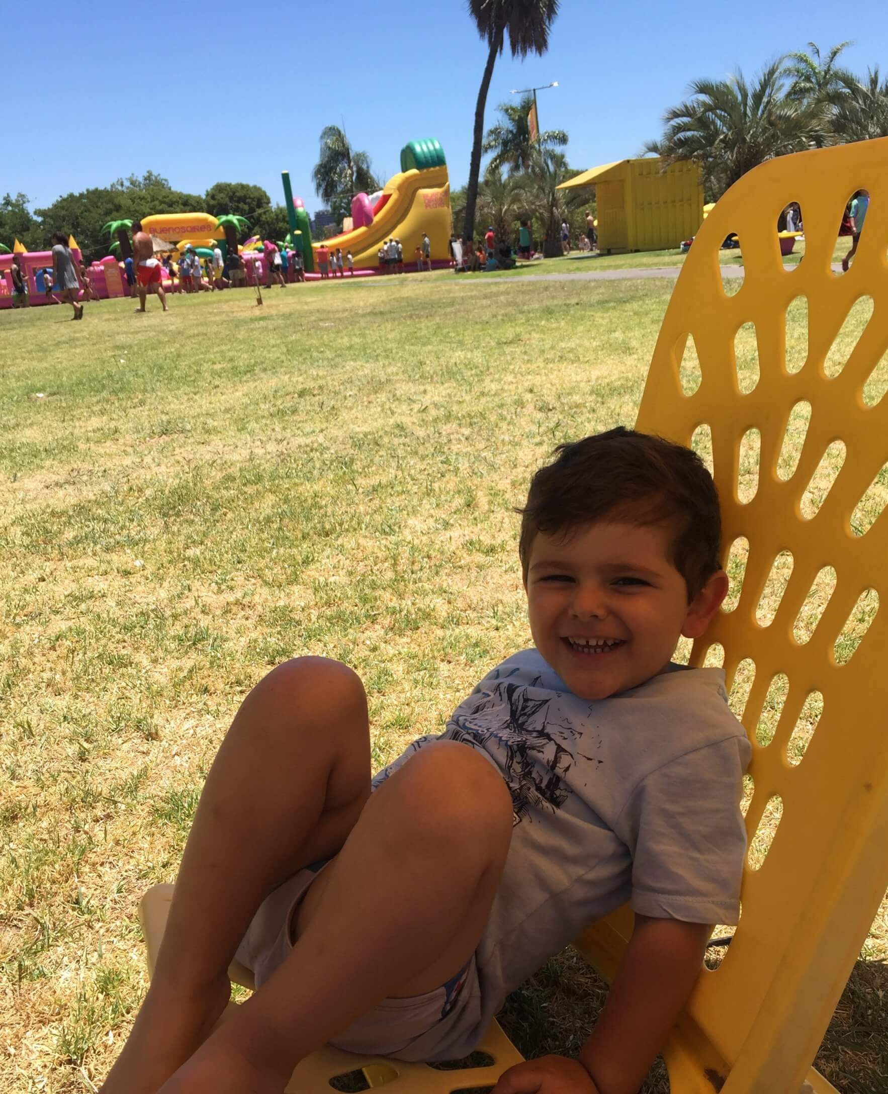Un niño sonriente está recostado en una silla amarilla de plástico sobre el césped. Al fondo se ven juegos inflables, palmeras y varias personas en un parque.