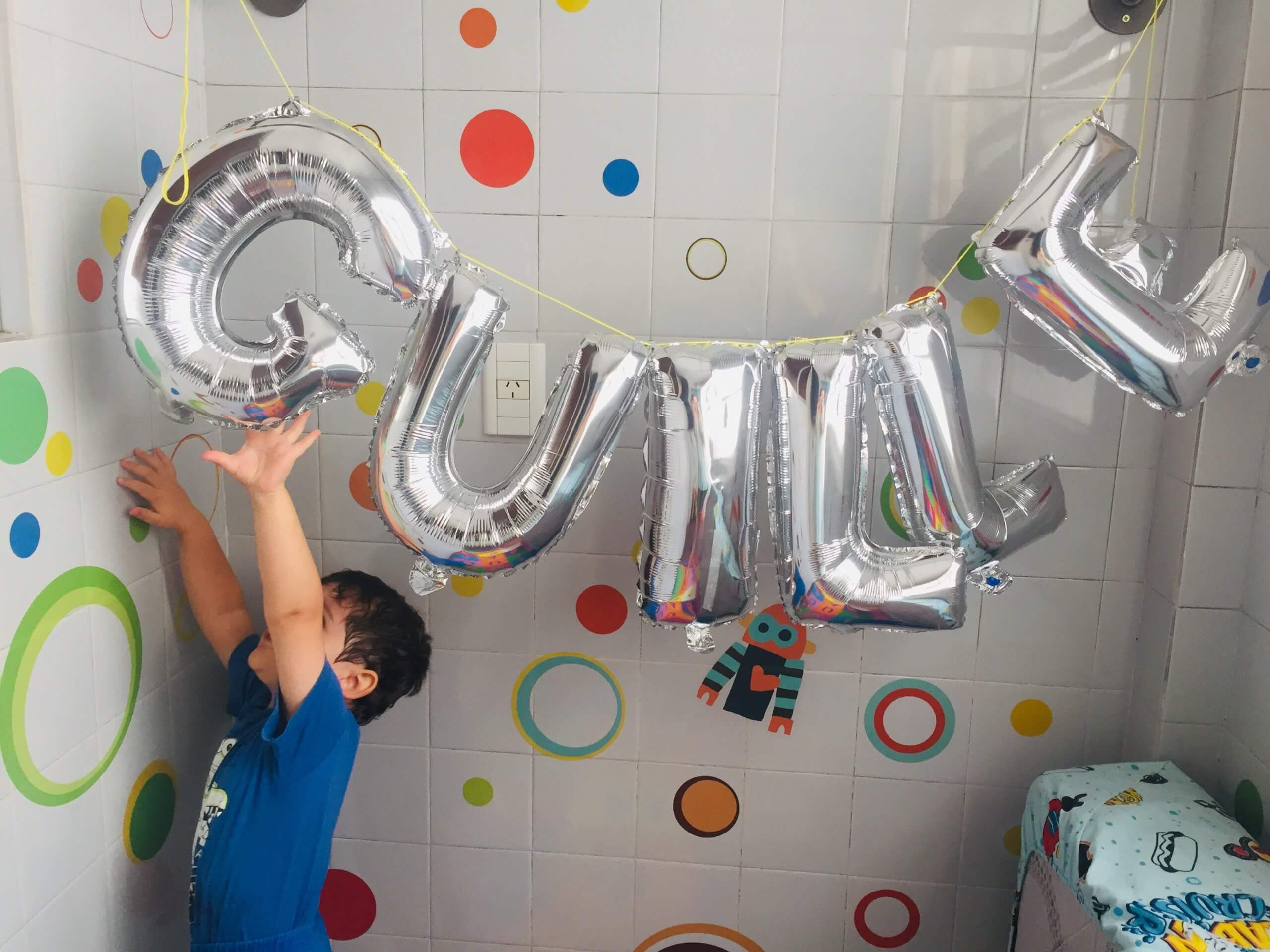 Un niño levanta los brazos hacia globos plateados con formas de letras, en un espacio decorado con círculos de colores.