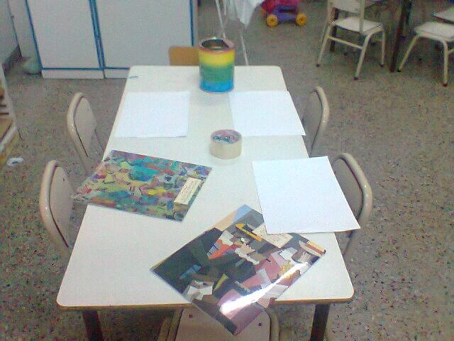 Mesa de aula con hojas blancas, una caja o tarro decorado en el centro y libros de imágenes abiertos o cerrados sobre la superficie.