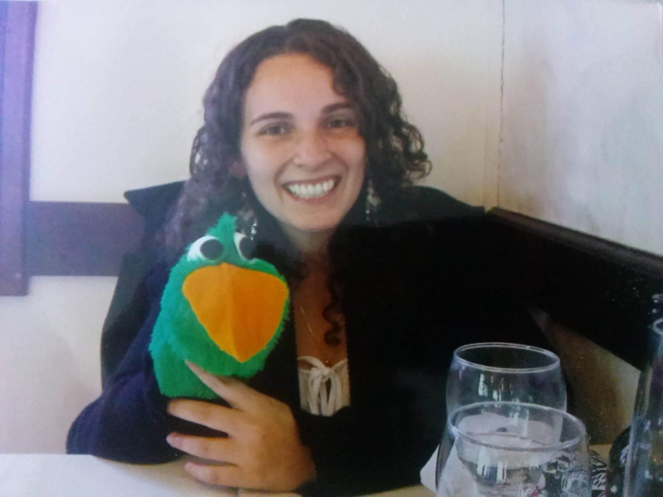 Fotografía de una persona sonriente sentada en una mesa y sosteniendo un títere verde con pico amarillo; delante hay vasos y al fondo se ve una pared clara.