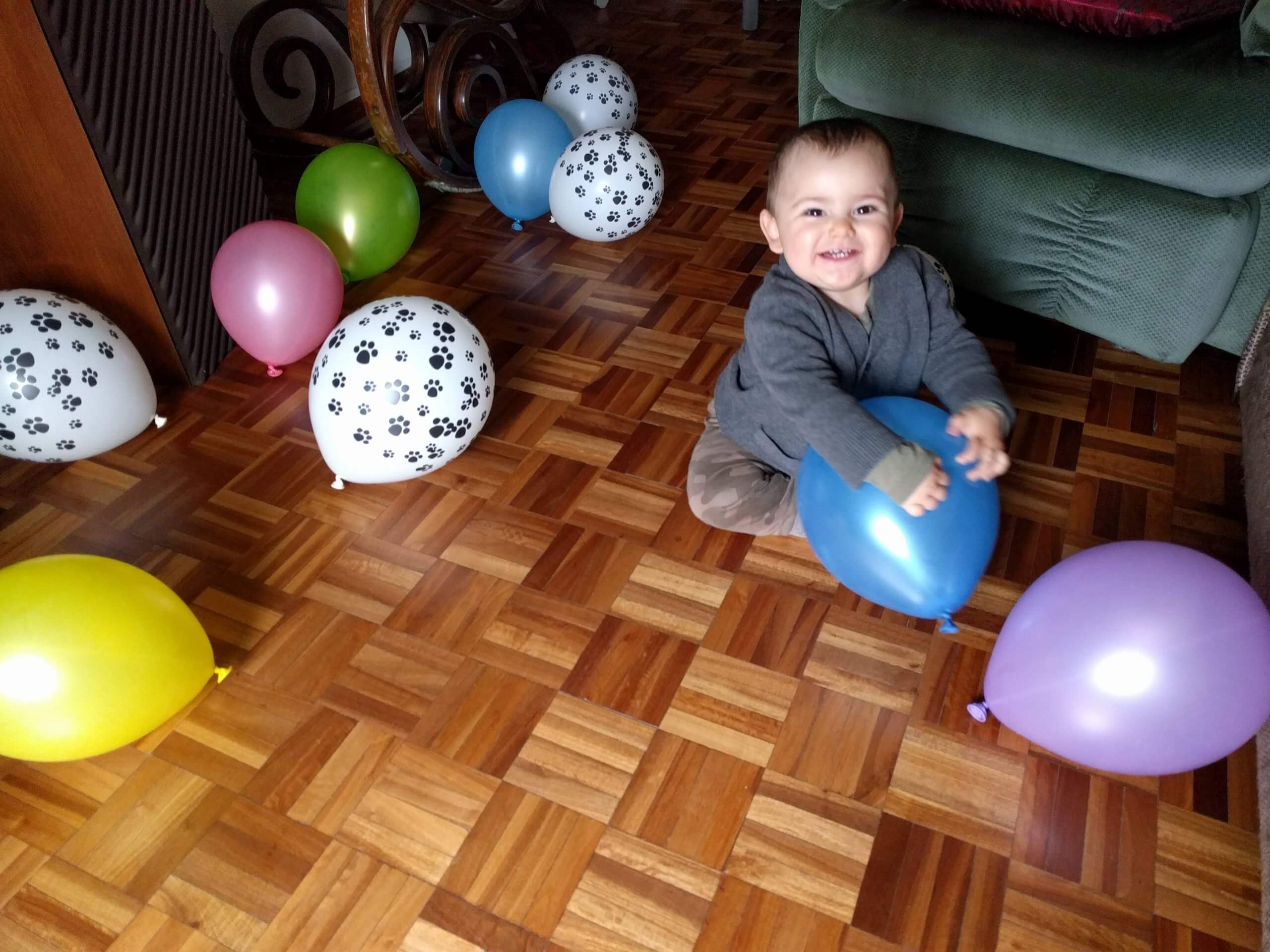 Bebé sentado entre globos en una sala Un bebé sonriente está sentado en el suelo de una sala, rodeado de globos de colores y globos blancos con estampas de huellas negras.