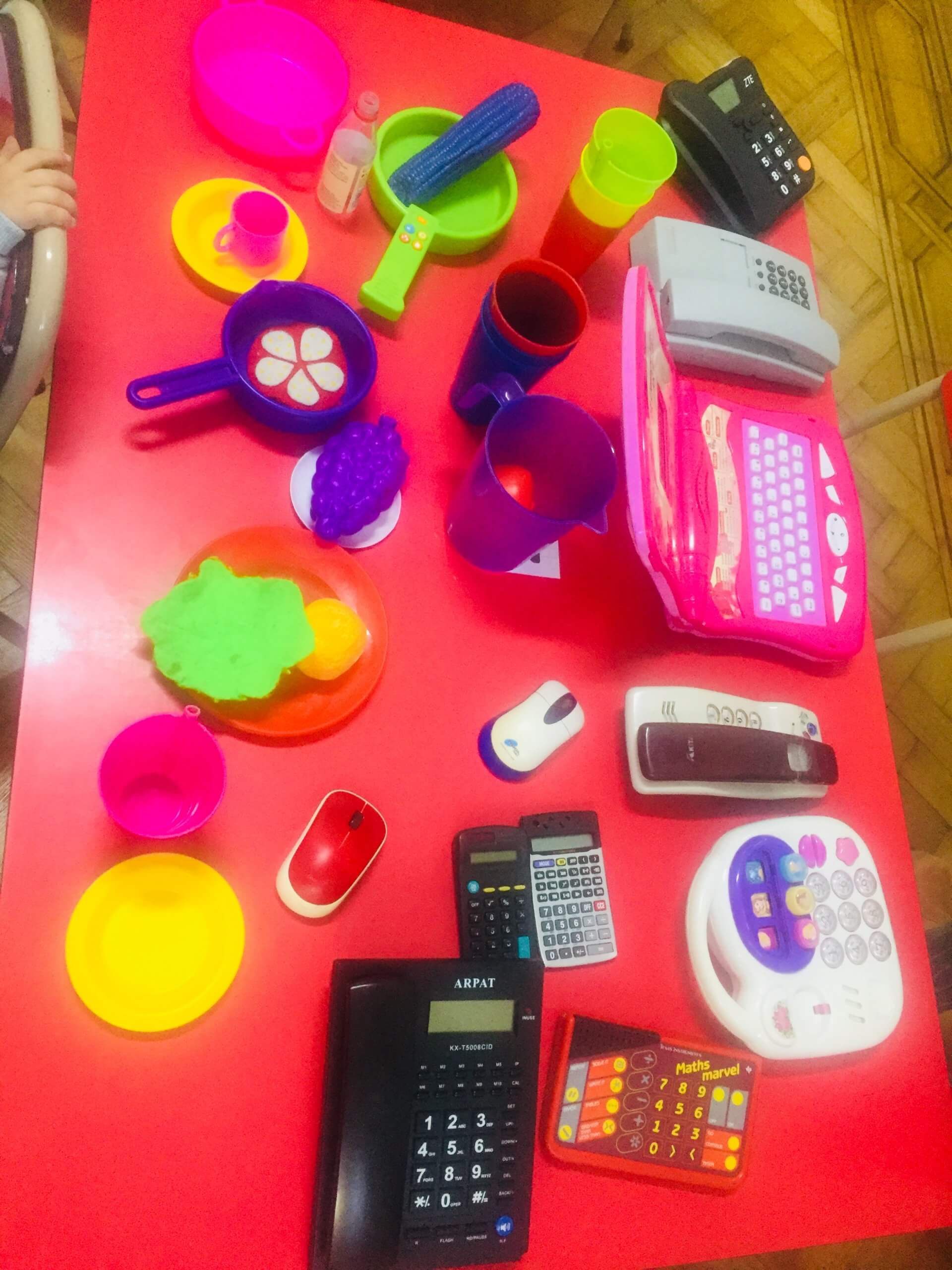Objetos cotidianos para el juego simbólico Mesa roja con teléfonos y calculadoras de juguete, platos, tazas, cucharas y otros utensilios de colores.