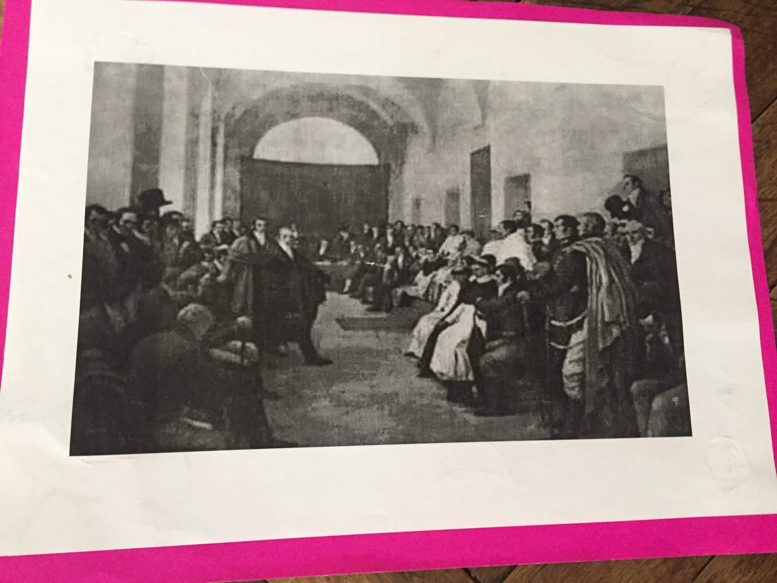 Imagen histórica de una reunión de personas en un salón Reproducción en blanco y negro de un interior amplio con muchas personas sentadas y de pie a ambos lados de un pasillo central. En el centro hay figuras principales de pie y al fondo se ve un gran arco o abertura.