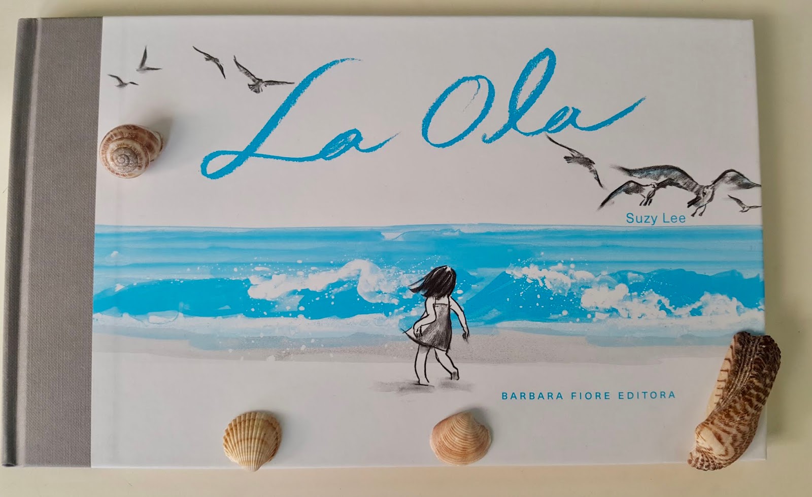 Tapa del libro "La Ola" decorada con caracoles Portada del libro La Ola, de Suzy Lee y Bárbara Fiore Editora, apoyada sobre una superficie clara y adornada con caracoles.