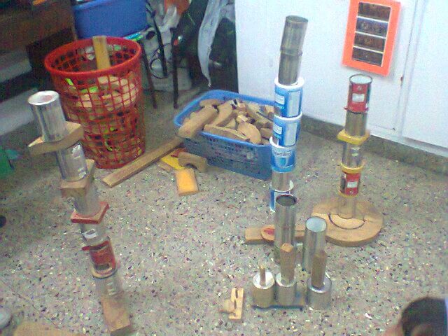 Varios montones de latas y piezas de cartón apiladas en el piso de un aula o sala de juego, junto a una canasta roja y otra azul con materiales sueltos.