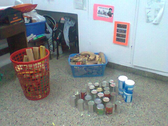 En un aula se ven grupos de latas apiladas en el piso, una canasta roja con piezas de cartón y una caja azul con bloques de madera contra la pared.