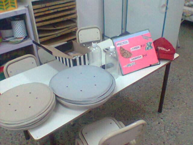 Mesa de aula preparada con varios platos de cartón apilados, botellas y un cartel rosado con opciones de comida para una actividad de pizzería.