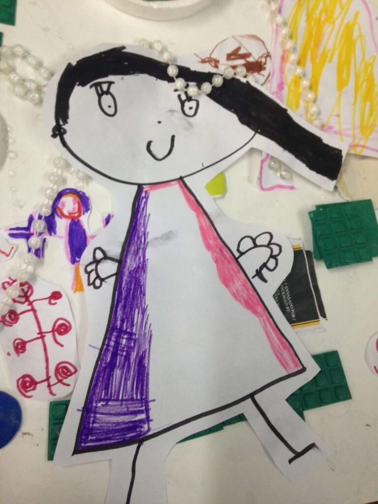 Un autorretrato infantil recortado en papel, con cabello negro y vestido rosa y violeta, junto a dibujos y materiales de aula.