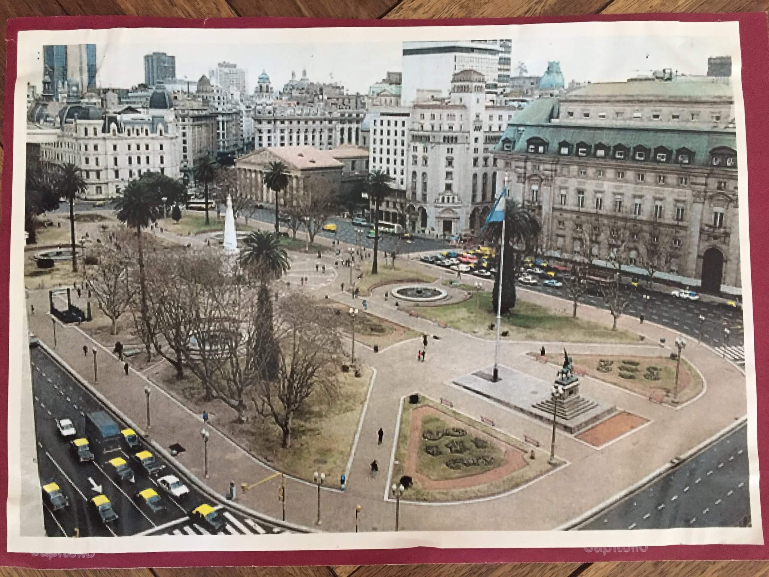 Plaza de Mayo vista desde arriba, con tránsito y edificios históricos Vista elevada de una plaza urbana amplia con senderos, monumentos, árboles y varios edificios históricos alrededor. Se ven autos circulando por las calles perimetrales.