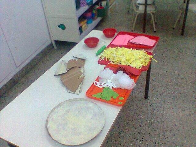 Mesa con bandejas de material didáctico que imita ingredientes para pizza, como queso rallado de papel, rodajas y piezas de cartón, junto a una base circular.