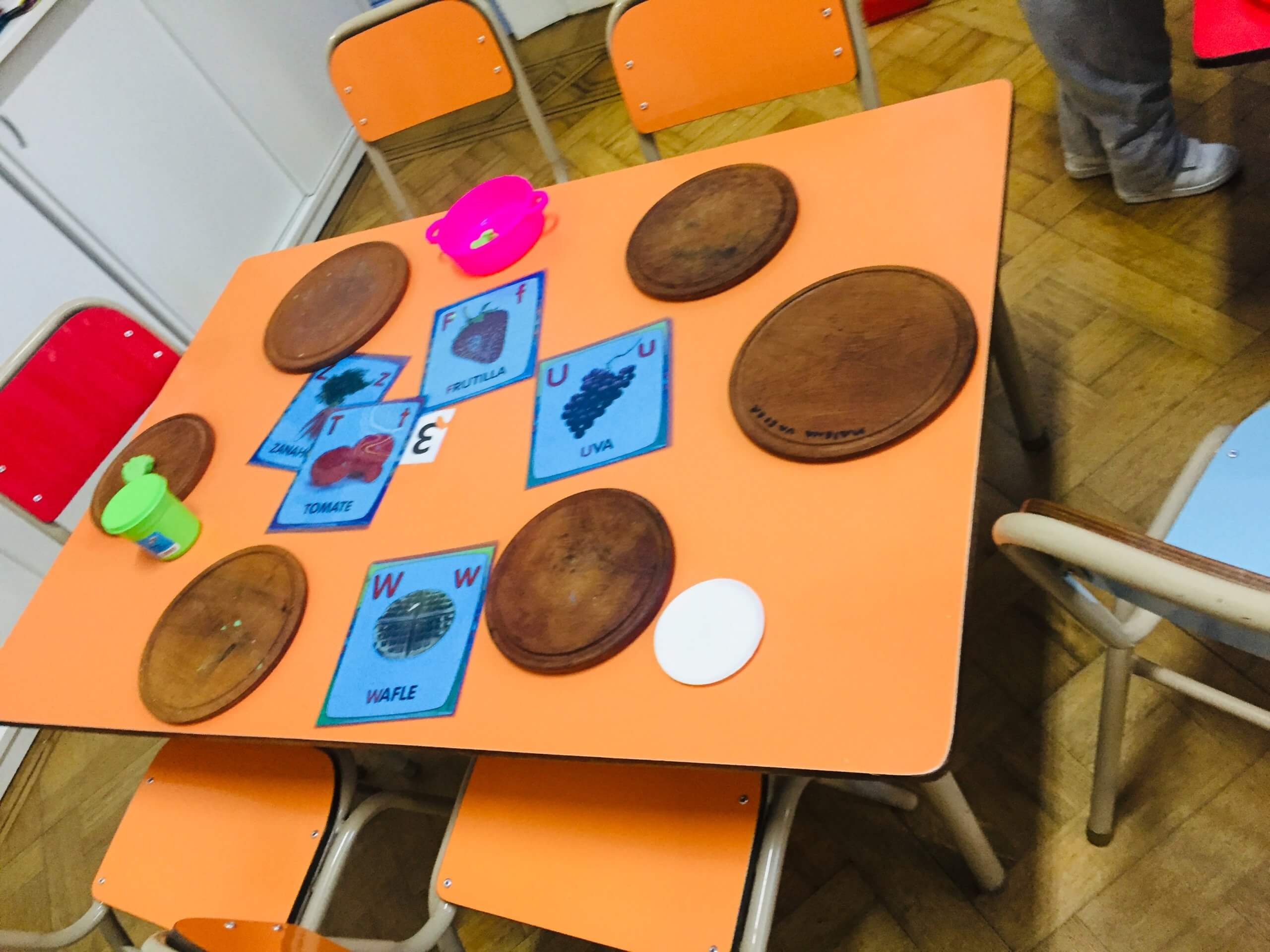 Tarjetas y materiales para la multitarea Mesa de la sala con tarjetas de frutas, bandejas y objetos de juego, junto a sillas de colores.