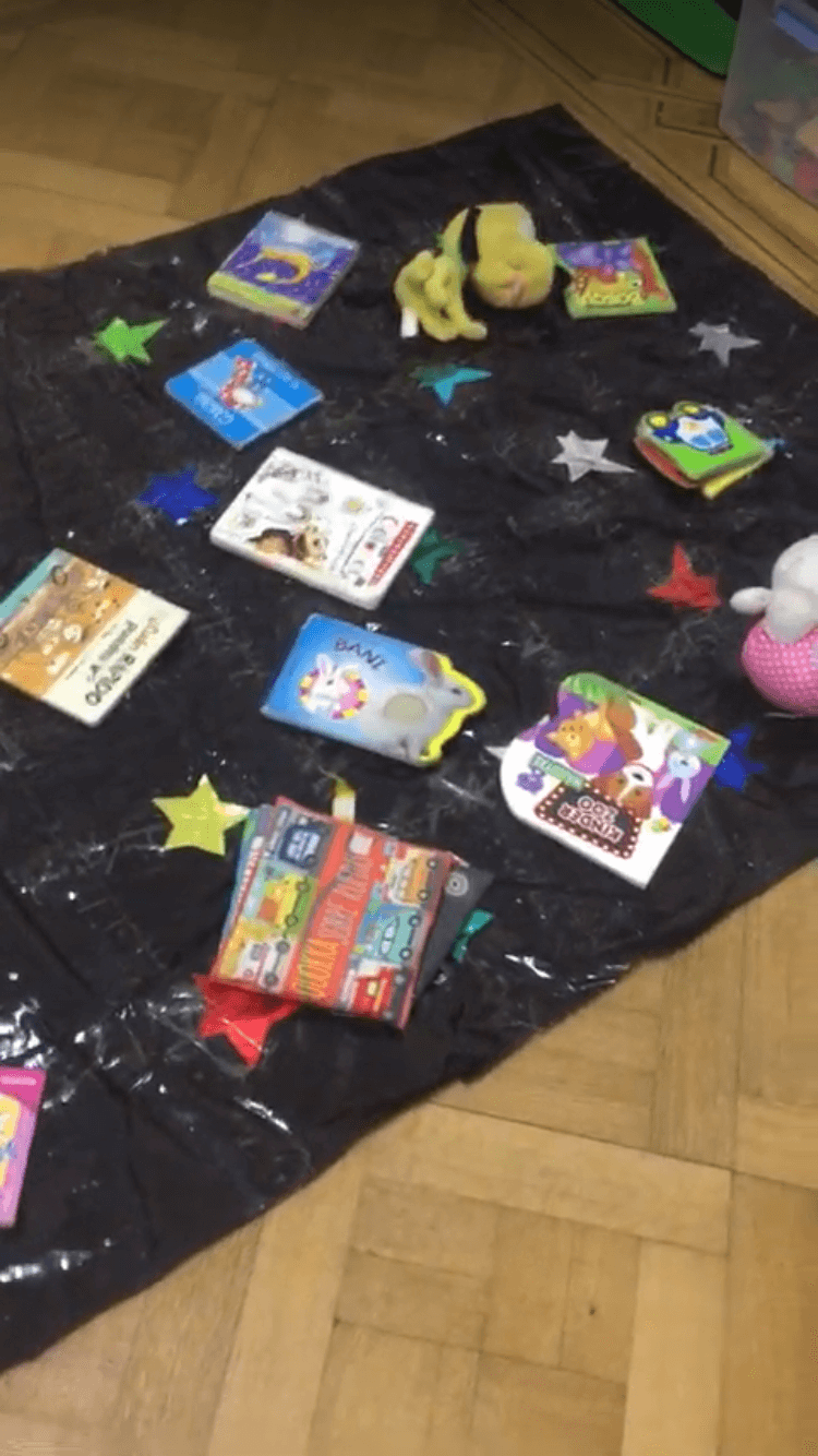Libros disponibles en un sector de lectura Libros abiertos y cerrados dispuestos sobre una tela negra con estrellas y almohadones en el piso.