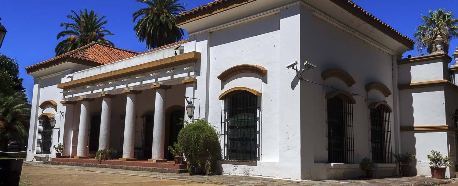 Casa histórica blanca con columnas y techo de tejas Edificio de una planta pintado de blanco, con columnas al frente, rejas en las ventanas y techo de tejas rojas. Alrededor hay palmeras y un cielo despejado.