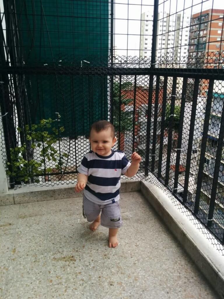 Exploración del movimiento en la primera infancia Un niño camina descalzo sobre el piso de una terraza con barandas y malla, con edificios al fondo.
