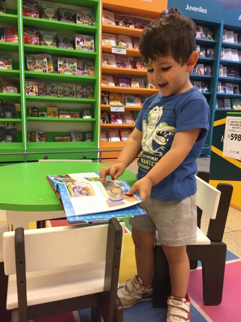 Lectura de libros para los 2 años Un niño pequeño hojea un libro ilustrado sobre una mesa verde junto a estanterías de libros infantiles.