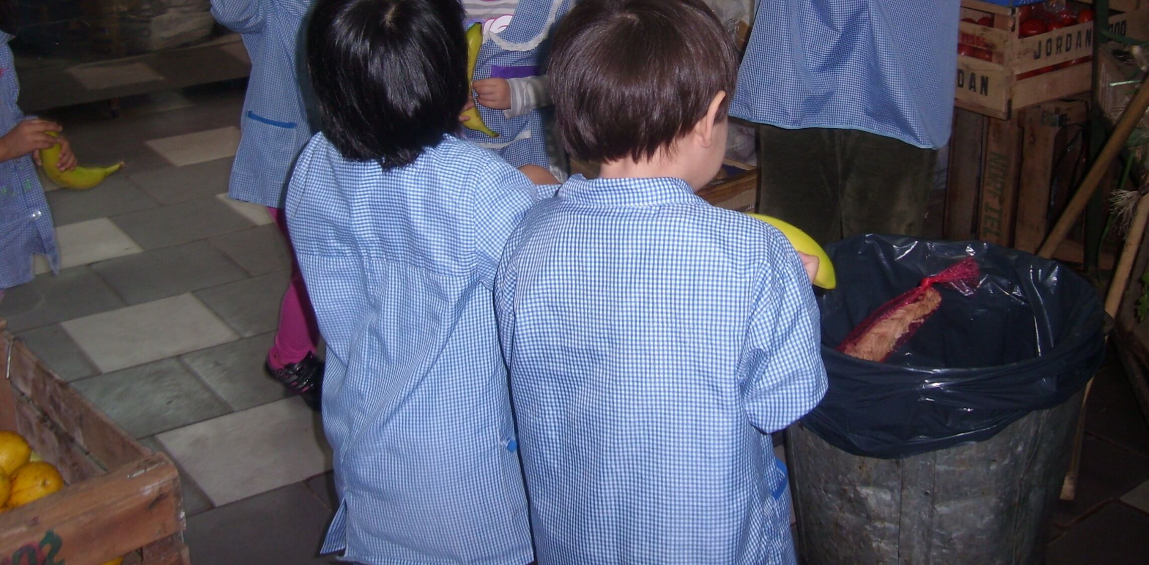 Reciclamos en el jardín: exploración de los residuos Varios niños con guardapolvo azul están de espaldas junto a un cesto con bolsa negra, en un espacio interior con cajones de verduras y piso de baldosas.