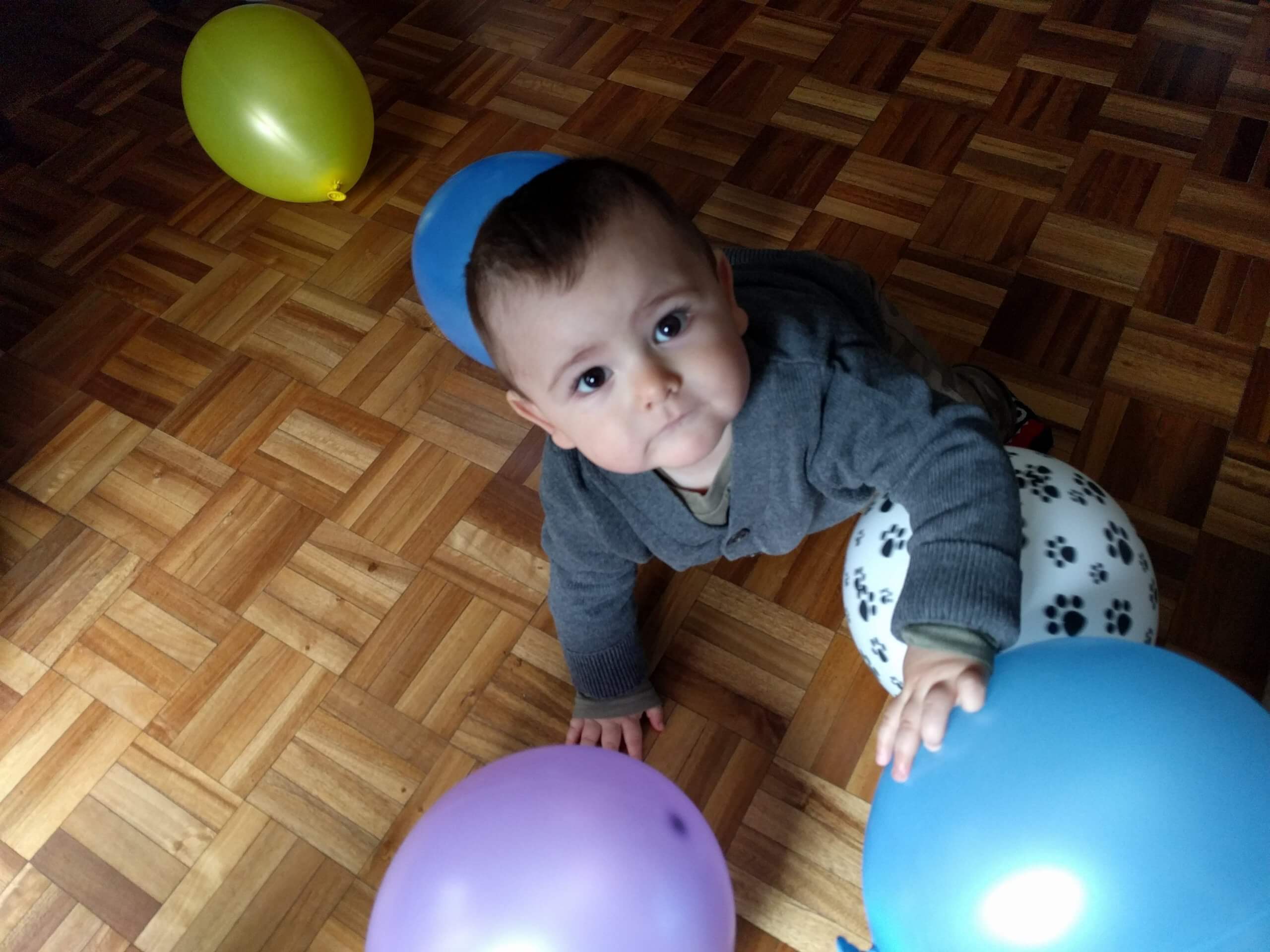 Bebé jugando con globos sobre piso de parquet Un bebé gatea sobre un piso de parquet y toca globos de colores, con uno azul en primer plano y otros globos detrás.