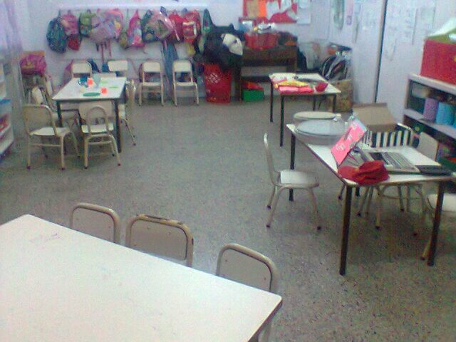 Vista general de un aula con varias mesas y sillas pequeñas, estantes con materiales y paredes decoradas, sin personas presentes.