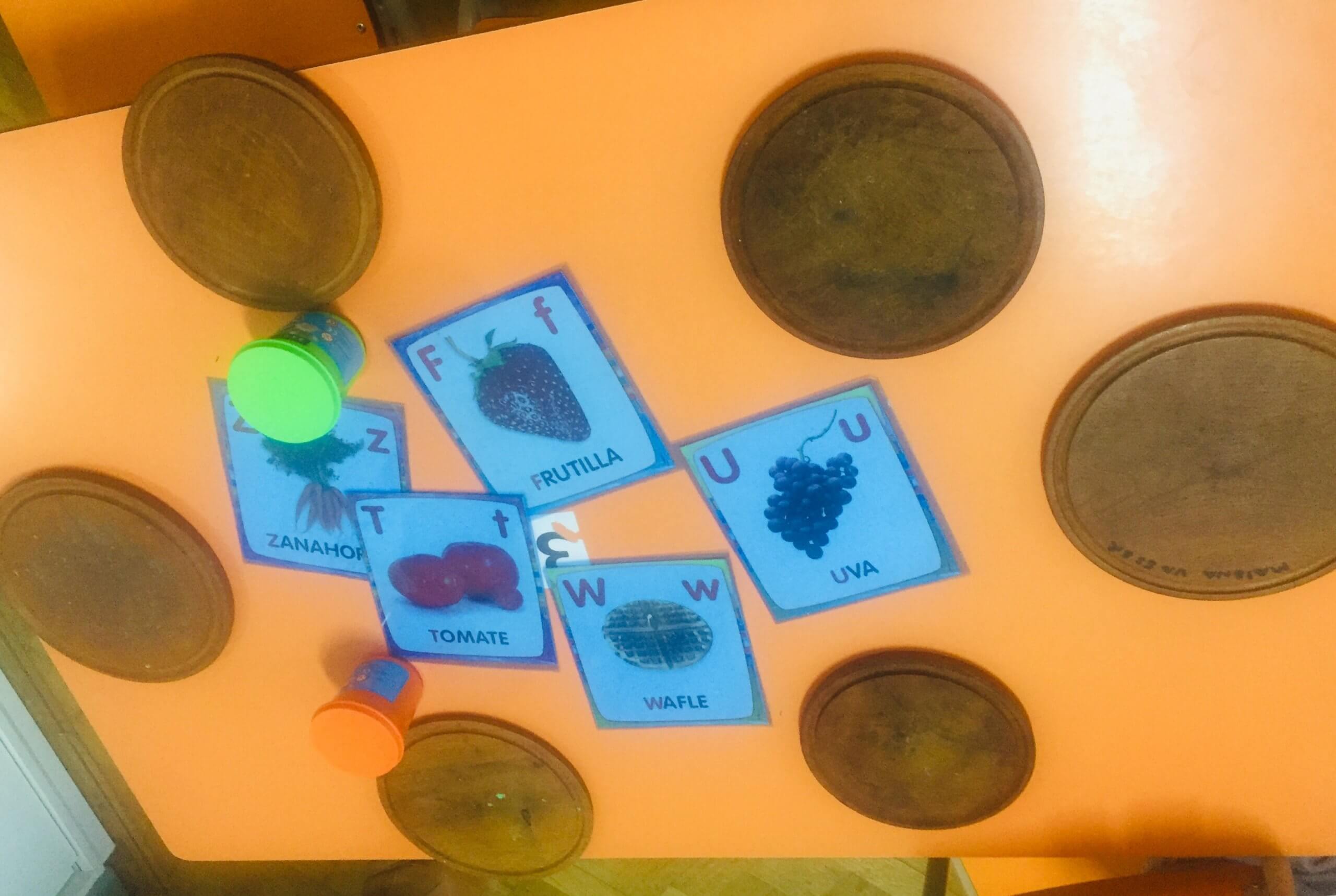 Juegos de lectura y exploración en una mesa de la sala Mesa naranja con bandejas circulares y tarjetas de frutas ilustradas y escritas, junto a objetos de juego.