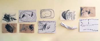 Dibujos y trazos infantiles expuestos en la pared Pequeños dibujos con trazos negros, formas circulares y representaciones pegados a una pared clara.