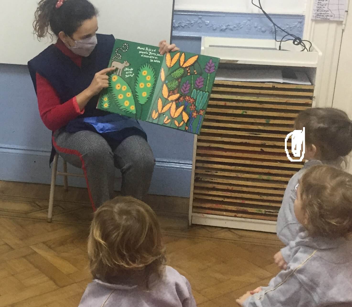Una docente con barbijo muestra un libro ilustrado abierto a niños pequeños sentados en el piso de madera del aula.