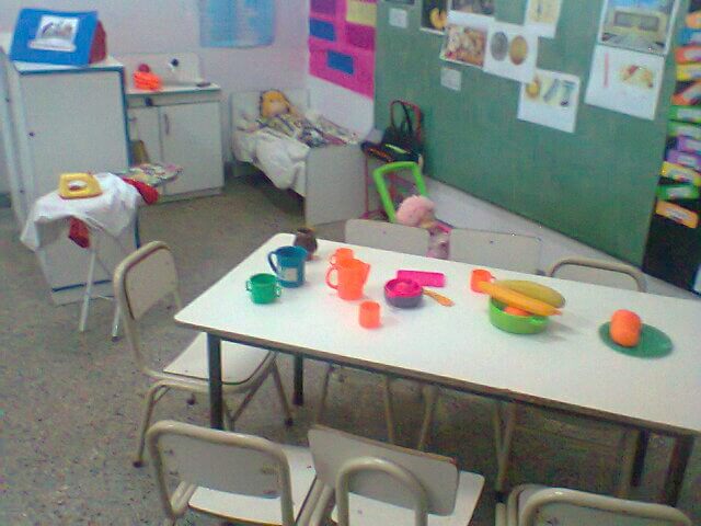 Mesa de aula con tazas, recipientes y utensilios de plástico de colores, frente a una pizarra verde con imágenes y dibujos pegados.