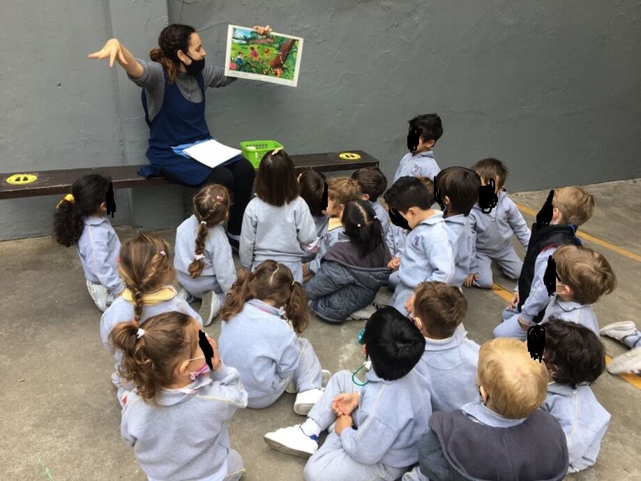 Una docente muestra una ilustración de un cuento a un grupo de niñas y niños sentados en el patio del jardín.