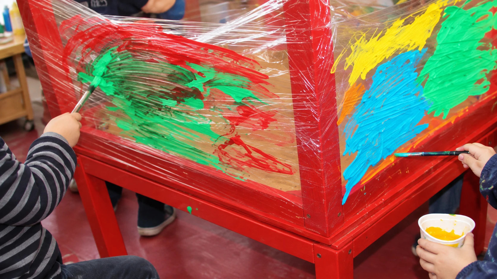 Pintar sobre una estructura cubierta con material transparente Recreación fotográfica de manos infantiles pintando con témperas sobre una estructura roja cubierta con material transparente.