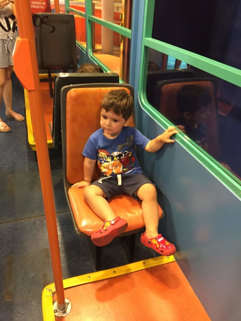 Un niño sentado en un asiento naranja dentro de una estructura de juego, junto a paneles de colores; imagen de la propuesta de viaje para sala de 2.
